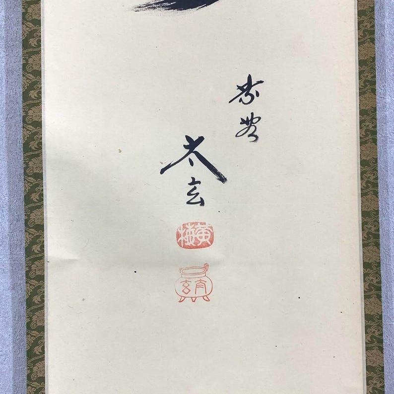 May include: A vertical scroll with Japanese calligraphy in black ink and red stamps. The calligraphy is on a cream-colored background, with a green and gold patterned border on the sides. The top of the scroll has a black ink brushstroke.