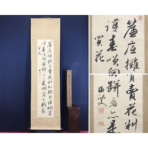 Op de afbeelding: Een antieke Chinese kalligrafierol met zwarte inkttekens op een crèmekleurige achtergrond. De rol hangt tegen een donkerblauwe muur. Een houten standaard en een houten blok met tekst zijn ook aanwezig.