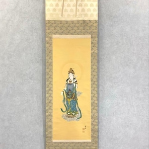 Op de afbeelding: Een traditioneel Japans rolschilderij met een figuur in blauwe en groene gewaden, tegen een gele achtergrond met een halo. De rol heeft decoratieve patronen en is opgehangen aan een zwarte ketting.