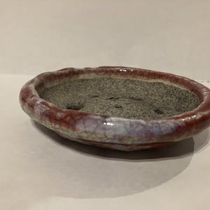 以下が含まれることがあります： 楕円形のセラミック盆栽鉢。縁は深い赤色で、ひび割れのある釉薬で、薄紫と白に変化しています。内側は質感のあるグレーで、小さな木の植え付けに適しています。