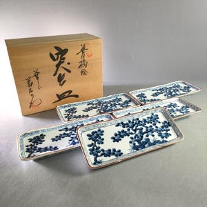 May include: Set of six rectangular ceramic serving dishes with a blue floral design on a white background. Each dish has a brown rim and a decorative blue border. A wooden box with Japanese calligraphy is in the background.