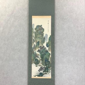 Könnte beinhalten: Vertikale asiatisch anmutende Rollbildmalerei mit einer Landschaft mit Bergen und Bäumen in Grün-, Blau- und Weißtönen. Das Kunstwerk ist auf einem grünen Stoffrücken mit einem dekorativen Muster montiert. Die Schriftrolle enthält Text in der oberen rechten Ecke.