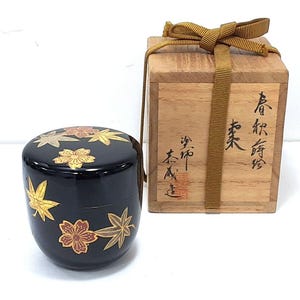 May include: A black, lacquered container with a lid, decorated with gold and red floral and leaf designs. The container is next to a wooden box tied with a gold ribbon. Japanese characters are visible on the box.