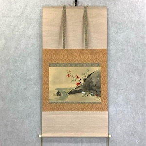 May include: A Japanese scroll painting with a landscape scene. The artwork shows a river, a rock formation, and flowering branches in green, red, and grey. The scroll is made of fabric and paper, with a wooden roller at the bottom.