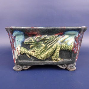 May include: Rectangular ceramic planter with a detailed green dragon relief against a black background. The planter has a dark blue, red, and white mottled glaze on the edges and sits on four small feet.