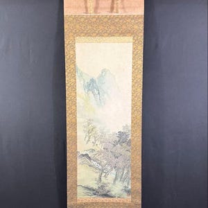 May include: An Asian scroll painting showing a mountain landscape with trees and a stream. The artwork uses muted colours, focusing on the natural scenery. The scroll is framed with decorative borders and has a wooden dowel at the bottom.