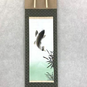 May include: Japanese hanging scroll with a carp fish illustration. The artwork depicts a carp swimming upwards against a light green and white backdrop. The scroll has a brown and green border with gold detailing.