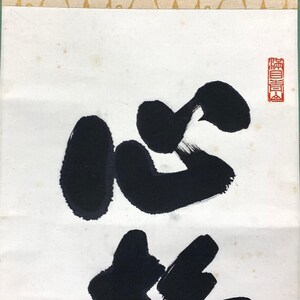 Puede incluir: Primer plano de una obra de caligraf&iacute;a japonesa tradicional. Gruesos trazos negros forman grandes caracteres sobre un fondo blanco. Un sello rojo est&aacute; en la esquina superior derecha. El papel tiene un aspecto texturizado.