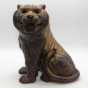 May include: A detailed, seated tiger sculpture with a fierce expression. The statue is primarily brown with lighter tan highlights, showcasing intricate carved details. The tiger's mouth is open, revealing teeth, and its pose is alert and ready.