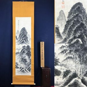 May include: Two Asian landscape paintings. One is a vertical scroll with a mountain scene in black and white ink on a cream background. The other is a larger painting with a similar mountain scene. A wooden box is in the foreground.