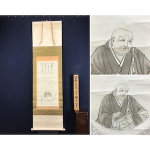 May include: An antique Japanese scroll painting with calligraphy and a detailed portrait of a monk. The scroll is cream-colored with a gold and green border. A wooden sign with Japanese text is next to the scroll.