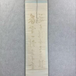 May include: A vertical scroll with Japanese calligraphy in faded gold ink. The scroll is cream-colored with a black rod at the bottom and a light blue top. The calligraphy is in a traditional style.