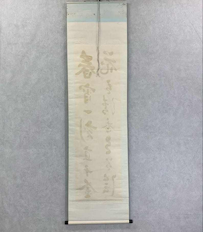 May include: A vertical scroll with Japanese calligraphy in faded gold ink. The scroll is cream-colored with a black rod at the bottom and a light blue top. The calligraphy is in a traditional style.