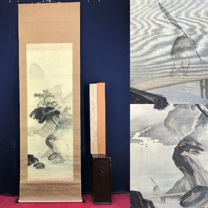 Könnte beinhalten: Traditionelle japanische Rollbildmalerei, die eine Landschaft mit Bergen, Bäumen und einem Fluss darstellt. Das Kunstwerk ist hauptsächlich in Grau- und Schwarztönen gehalten, mit einem beigen Rand und Hintergrund. Neben der Rolle befindet sich eine Holzkiste mit Schlüsselloch.