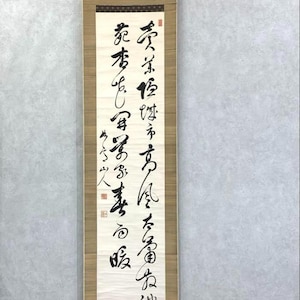 May include: A vertical Japanese scroll with calligraphy in black ink on a cream-colored background. The scroll is framed by a light brown wooden border and has a dark brown top and bottom. The text is written in vertical columns.