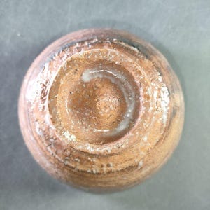 May include: A handmade ceramic bowl, viewed from the bottom. The bowl features a circular design with concentric rings and a central indentation. The color is a mix of brown and tan with white accents, showcasing a rustic aesthetic.