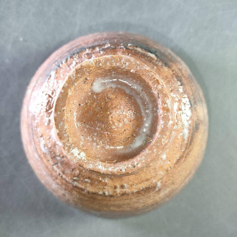 May include: A handmade ceramic bowl, viewed from the bottom. The bowl features a circular design with concentric rings and a central indentation. The color is a mix of brown and tan with white accents, showcasing a rustic aesthetic.