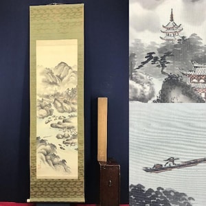 Op de afbeelding: Een traditioneel Japans rolschilderij dat een berglandschap met een rivier uitbeeldt. Het kunstwerk gebruikt grijstinten en wit, met een groene patroonrand. Een houten staaf en een kleine houten doos zijn ook zichtbaar.