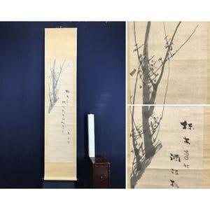 May include: A long, vertical Asian scroll painting featuring a black ink depiction of a tree branch with small blossoms. The scroll is cream-colored with black calligraphy. The background is a dark blue wall.