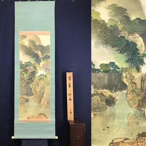 May include: A traditional Japanese scroll painting depicting a landscape with mountains, trees, and water. The artwork features muted colours and a serene atmosphere. A wooden box with Japanese characters is next to the scroll.