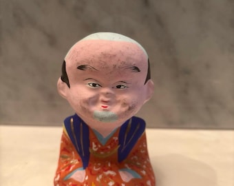May include: A small, hand-painted figurine of a man with a bald head and a pink face. The figure wears a red and orange robe with blue accents. The man has black hair, eyebrows, and a small blue goatee.