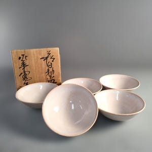May include: A collection of five off-white ceramic bowls with a pale pink rim. The bowls are arranged in a semi-circle, with a wooden box in the background. The box features Japanese calligraphy.