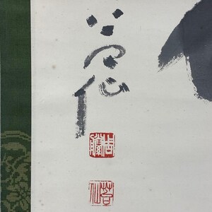 May include: Close-up of a traditional Asian artwork featuring black ink calligraphy on white paper. The artwork includes several characters and red stamps, with a green patterned border on the left side.
