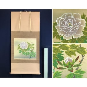 Op de afbeelding: Een verticale hangende rol met een schildering van een witte pioenroos met groene bladeren op een lichte achtergrond. De rol heeft beige stoffen randen en twee ophanglinten aan de bovenkant. Detailopnames tonen de bloem en een bloemknop.