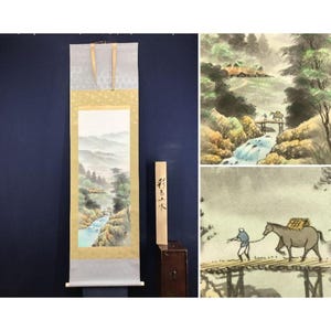 Könnte beinhalten: Ein japanisches Rollbild, das eine Berglandschaft mit einem Fluss und einer Brücke zeigt. Das Kunstwerk weist gedämpfte Farben und einen traditionellen Stil auf. Ein Holzschild mit japanischen Schriftzeichen steht neben der Schriftrolle.