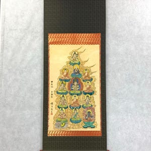May include: A vertical Japanese scroll painting featuring a detailed illustration of multiple seated figures with halos. The artwork has a cream-colored background with a red and gold border, and is framed by a dark green textured fabric.