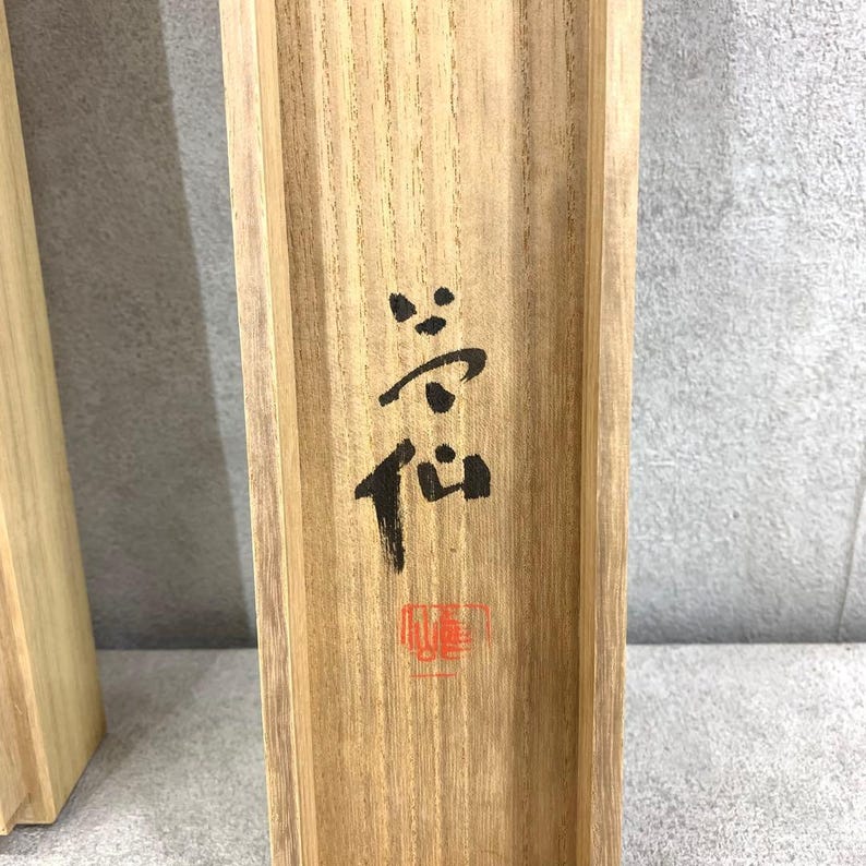 May include: Close-up of a wooden box with Japanese calligraphy in black ink. The characters are vertically oriented and include a red stamp at the bottom. The wood has a natural grain and a light brown color.
