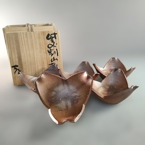 以下が含まれることがあります： ユニークな花びらのようなデザインの、茶色の陶器のボウル4個セット。 ボウルは灰色の表面に配置され、背景には木製の箱があります。 箱には日本語の文字が書かれています。