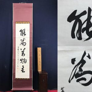 以下が含まれることがあります： 白い紙に黒インクで書かれた書道が施された、縦長の日本の巻物。巻物には、バーガンディ色の縁と、金と茶色の装飾的な帯があります。木箱と小さな書道作品も存在します。