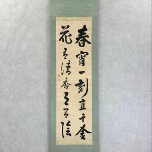 May include: A vertical scroll with Japanese calligraphy in black ink on cream-colored paper. The scroll is framed by a light green fabric with a decorative top and bottom. The text is in a traditional East Asian script.
