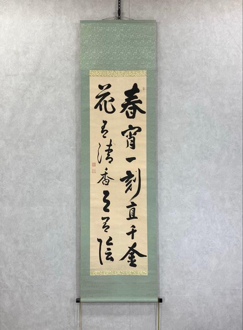 May include: A vertical scroll with Japanese calligraphy in black ink on cream-colored paper. The scroll is framed by a light green fabric with a decorative top and bottom. The text is in a traditional East Asian script.