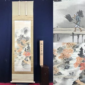 Op de afbeelding: Een traditioneel Japans rolschilderij dat een landschap met bergen, bomen en een brug uitbeeldt. Het kunstwerk heeft gedempte kleuren, met tinten rood, oranje en groen. Een houten kist met Japanse tekst staat naast de rol.