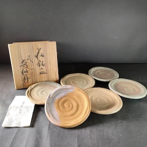 Könnte beinhalten: Ein Set aus sechs Keramikplatten mit Spiralmuster und einer Holzbox. Die Platten haben eine Mischung aus Erdtönen, darunter Braun, Beige und ein Hauch von Blau. Die Holzbox hat japanische Schriftzeichen.