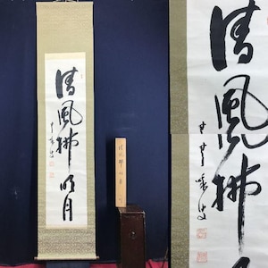 May include: A vertical scroll with black calligraphy on white paper, mounted on a beige background. The calligraphy features bold brushstrokes and vertical text. A small wooden block with text stands to the side. The background is a dark blue.