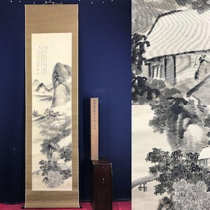May include: An Asian landscape painting on a scroll, depicting mountains, trees, and a building in shades of grey and black. The scroll is mounted on a tan backing, with a wooden box and a wooden stick. The artwork is a traditional style.
