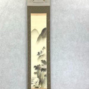 May include: A vertical Japanese scroll painting showing a misty mountain landscape. The artwork uses muted tones of grey, beige, and brown. The scroll is framed with a patterned border and has wooden dowels at the top and bottom.