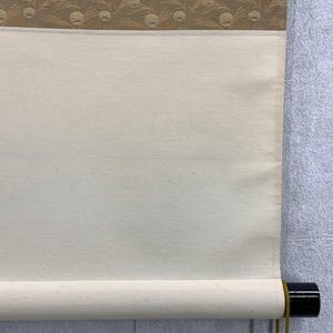 May include: A close-up of a traditional Japanese scroll. The scroll features a cream-colored paper with a decorative border of tan and gold circular patterns. The scroll is rolled up with a black wooden rod at the bottom.