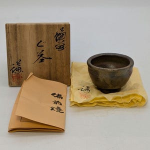 May include: A small, dark brown ceramic bowl with a rounded shape, on a yellow cloth. A wooden box with Japanese characters and a paper envelope are also present. The bowl has a textured surface.