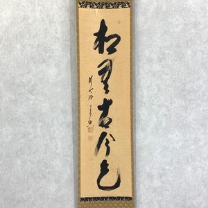 May include: A rectangular, vertical scroll with Japanese calligraphy in black ink. The scroll has a tan background with a decorative border at the top and bottom. The calligraphy is written vertically, with several characters.