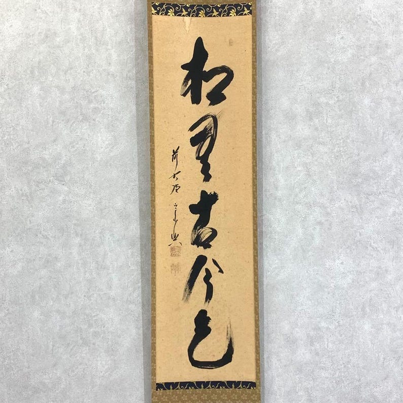 May include: A rectangular, vertical scroll with Japanese calligraphy in black ink. The scroll has a tan background with a decorative border at the top and bottom. The calligraphy is written vertically, with several characters.