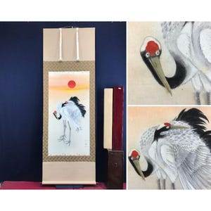 May include: A Japanese hanging scroll featuring a painting of two white cranes with red crowns and black wing tips standing against a soft orange sunset with a red sun. The scroll has a beige border with a patterned fabric frame and white hanging cords.