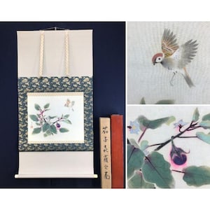 May include: A Japanese scroll painting with a bird and botanical illustration. The artwork features a bird in flight and a branch with purple fruit and green leaves. The scroll has a blue floral border and is displayed with wooden blocks.