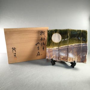 May include: A square ceramic plate with a landscape design featuring a white moon, green and brown tones, and a wooden box with Japanese calligraphy. The plate is displayed on a black stand.