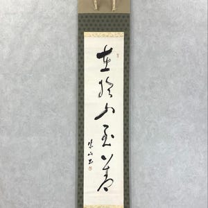 May include: A vertical Japanese scroll with calligraphy in black ink on a white background. The scroll has a tan top and bottom, with a patterned green border. The calligraphy is in a traditional style.