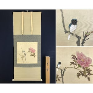 Op de afbeelding: Een traditioneel Japans rolschilderij met een roze pioenroos en een kleine vogel die op een tak zit. De rol heeft een lichtgroene en grijze achtergrond met een houten opbergdoos.