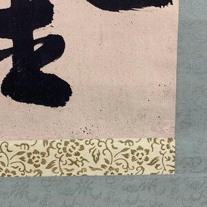 May include: Close-up of a traditional Asian scroll painting. The artwork features bold black calligraphy on a light pink background. The scroll is bordered with a decorative floral pattern and a light blue fabric.